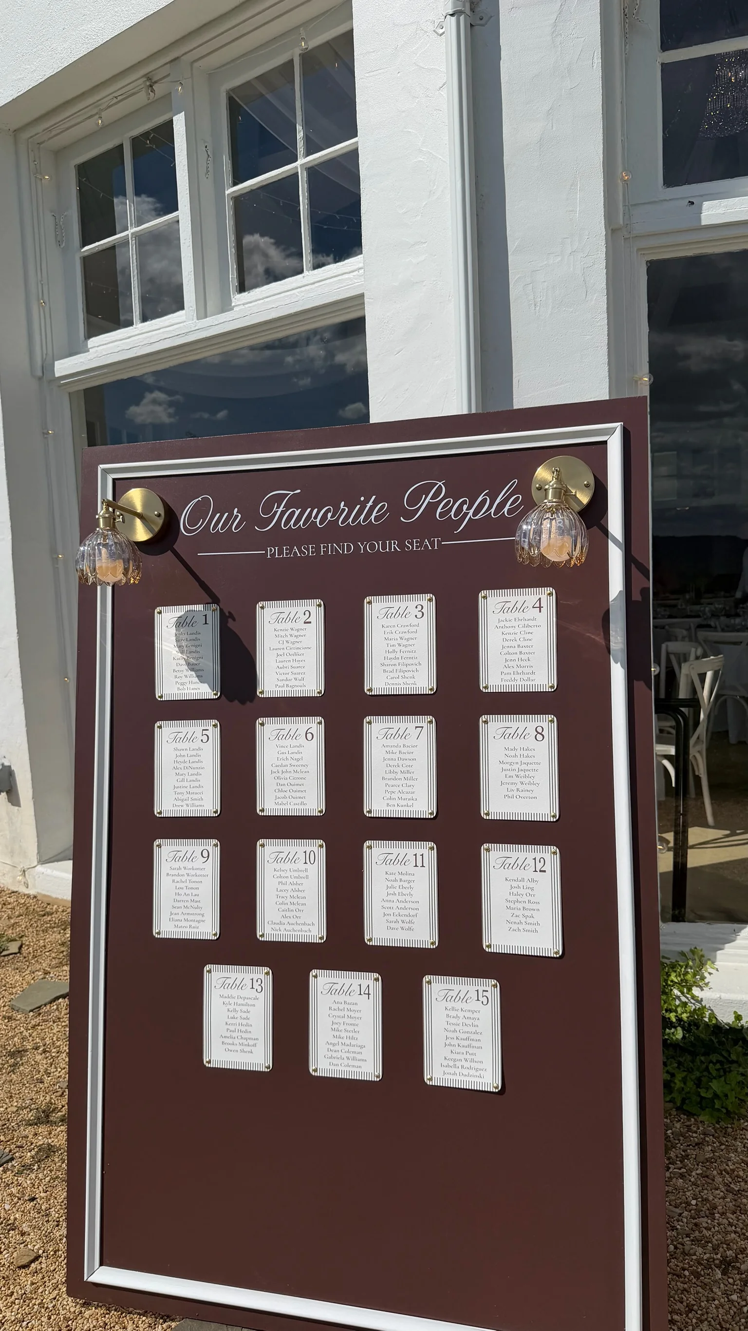 Brown framed Our Favorite People seating chart with rows of white table cards and brass lights